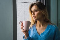 Стомлена молода жінка п'є каву у вікні — стокове фото