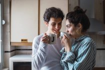 Счастливая молодая пара стоит с двумя чашками и улыбается на кухне утром — стоковое фото