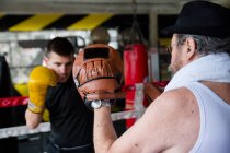 Вид збоку боксерський чоловік ударяє рукавички тренера в спортзалі . — стокове фото