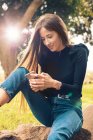 Молодая улыбающаяся женщина сидит на камне и использует смартфон в парке — стоковое фото