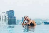 Mulher asiática relaxante na piscina com arranha-céus no fundo — Fotografia de Stock