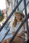 Giovane donna bionda in cappello e vestito estivo seduta su gradini fuori a guardare lontano in sogno ad occhi aperti — Foto stock