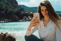 Woman taking selfie with smartphone at rocky seaside — Stock Photo