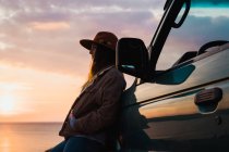 Mulher pensativa apoiando-se no carro na costa do mar ao pôr do sol — Fotografia de Stock