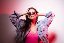 Pretty young pink dressed woman in sunglasses standing at white wall with hands in hair — Stock Photo
