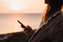 Femme utilisant un smartphone tout en se tenant sur la côte au coucher du soleil — Photo de stock
