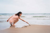 Menina adolescente agachamento e pintura com pau na areia na praia — Fotografia de Stock