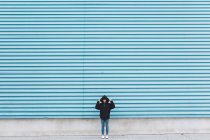 Hombre en chaqueta con capucha de pie contra la pared de metal azul - foto de stock