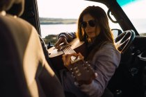 Attraente giovane donna che suona la chitarra e seduta in auto nella natura — Foto stock