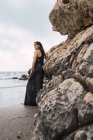 Модная женщина в черном платье, стоящая у скалы на пляже — стоковое фото