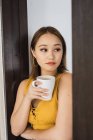 Задумчивая молодая женщина с чашкой опираясь на стену дома — стоковое фото