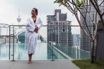 Woman in bathrobe standing at pool with modern buildings on background — Stock Photo