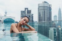 Portrait of Asian woman relaxing in pool with modern skyscrapers on background — Stock Photo
