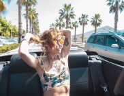 Mujer sonriente en gafas de sol y ropa de verano corta cabalgando en el asiento del pasajero en cabriolet con las manos en el aire en la carretera con palmeras alrededor - foto de stock