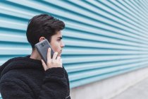 Молодой подросток, стоящий у металлической стены и разговаривающий на смартфоне на улице — стоковое фото