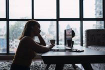 Frau mit Tasse mit Laptop am Tisch in moderner Wohnung — Stockfoto