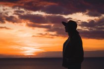 Mulher pensativa em chapéu de pé no mar ao pôr do sol — Fotografia de Stock