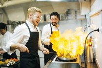 Heureux cuisinier faire une flamme dans la cuisine du restaurant avec collègue regarder — Photo de stock