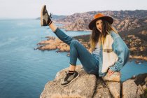 Портрет стильной веселой женщины в шляпе, сидящей на скале у океана — стоковое фото