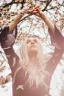 Young blonde woman standing at blooming tree with hands up — Stock Photo
