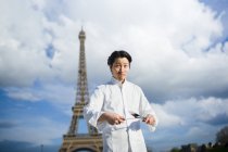 Portrait of smiling Japanese chef with knives standing in front of Eiffel Tower in Paris — Stock Photo