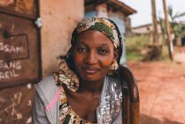 Kamerun - Afrika - 5. April 2018: lächelnde erwachsene afrikanische Frau, die in die Kamera schaut, während sie im Haus an der Dorfstraße sitzt — Stockfoto