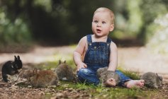 Милий маленький хлопчик в джинсовому одязі, сидячи з маленькими кроликами на землі в парку — стокове фото