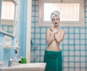 Jeune femme seins nus enveloppée dans des serviettes regardant la caméra dans la salle de bain bleue — Photo de stock