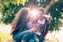 Смеющаяся молодая пара сидит со смартфоном в парке — стоковое фото