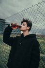 Junger schöner Mann trinkt aus Plastikflasche am Zaun — Stockfoto