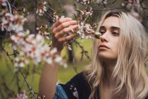 Junge blonde Frau steht am blühenden Baum und berührt Blüte — Stockfoto