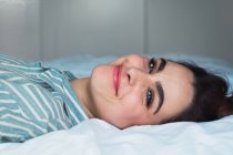 Fröhliche junge brünette Frau im Pyjama auf dem Bett liegend und in die Kamera schauend — Stockfoto