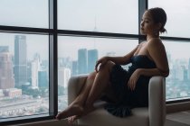 Jolie femme en robe noire relaxante dans un fauteuil dans un appartement avec vue sur la ville — Photo de stock
