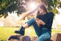 Junge lächelnde Frau sitzt auf einem Felsen und benutzt Smartphone im Park — Stockfoto