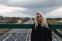 Thoughtful blonde woman standing at fence on bridge over road — Stock Photo