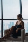 Jolie femme en robe noire relaxante dans un fauteuil dans un appartement avec vue sur la ville — Photo de stock