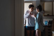 Jovem casal alegre de pé com xícaras de café na cozinha de manhã — Fotografia de Stock