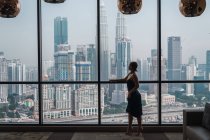 Mujer de pie en la ventana en el apartamento y mirando los rascacielos - foto de stock