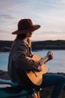 Woman sitting on car roof and playing guitar on coast at sunset — Stock Photo