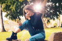 Молода жінка сміється сидить на камені і використовує смартфон у парку — стокове фото