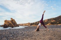 Femme sportive debout avec la jambe vers le haut et pratiquer le yoga sur le rivage à l'océan — Photo de stock