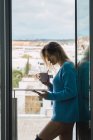 Sensual mujer joven en suéter sosteniendo taza de café y surf smartphone en la puerta del balcón - foto de stock