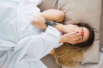 Verspielte junge Frau bedeckt Gesicht mit Händen im Bett — Stockfoto