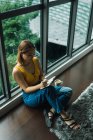 Жінка з чашкою, що пише в блокноті, сидячи на підлозі у вікні — стокове фото