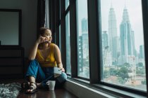 Donna con tazza e taccuino seduta sul pavimento alla finestra — Foto stock