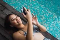 Улыбающаяся женщина со смартфоном расслабляется у бассейна — стоковое фото
