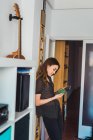 Frau benutzt Handy, während sie sich zu Hause an Wand lehnt — Stockfoto