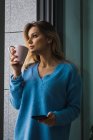 Mujer rubia pensativa sosteniendo teléfono inteligente y taza de café en la ventana - foto de stock