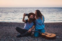 Casal elegante com guitarra na praia ao pôr do sol tomando selfie — Fotografia de Stock
