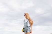 Blonde girl standing with penny board in front of cloudy sky — Stock Photo
